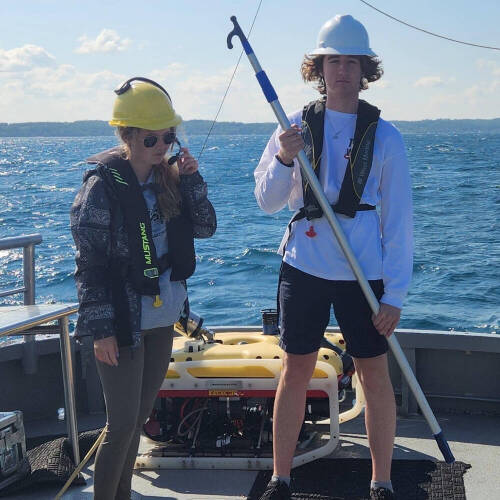Two individuals on a boat deck with a yellow underwater drone, wearing hard hats and life vests against a blue ocean backdrop.